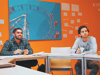 Zwei Kursteilnehmer in Wien sitzen im hellen Klassenraum und hören aktiv zu.