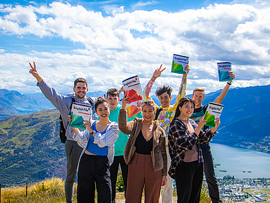 Sprachschüler halten ihre Englischbücher hoch mit Blick auf die Berge von Queenstown.