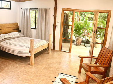 Helles Schlafzimmer im Iona Studio Sámara mit rustikalem Holzbett, großen Fenstern und Zugang zur begrünten Terrasse.