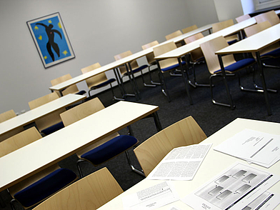 Moderner Klassenraum mit hellen Tischen und Stühlen, blauen Sitzpolstern und Unterrichtsmaterialien auf dem Tisch