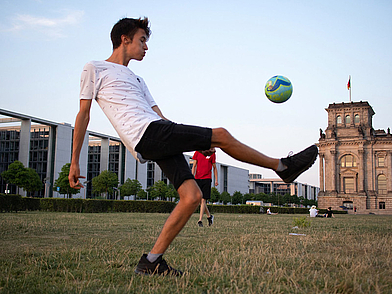 Jugendlicher spielt Fußball auf einer Wiese mit Blick auf das Reichstagsgebäude