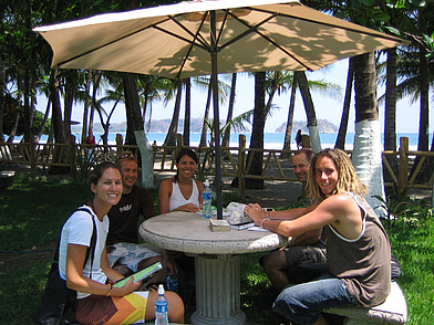 Gruppe internationaler Sprachschüler der Intercultura Sprachschule Sámara sitzt unter Palmen am Tisch mit Blick auf den Strand.